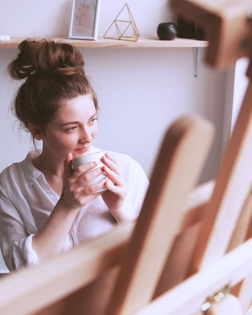 rapariga com bun no cabelo com uma caneca entre as mãos, de frente para um cavalete.