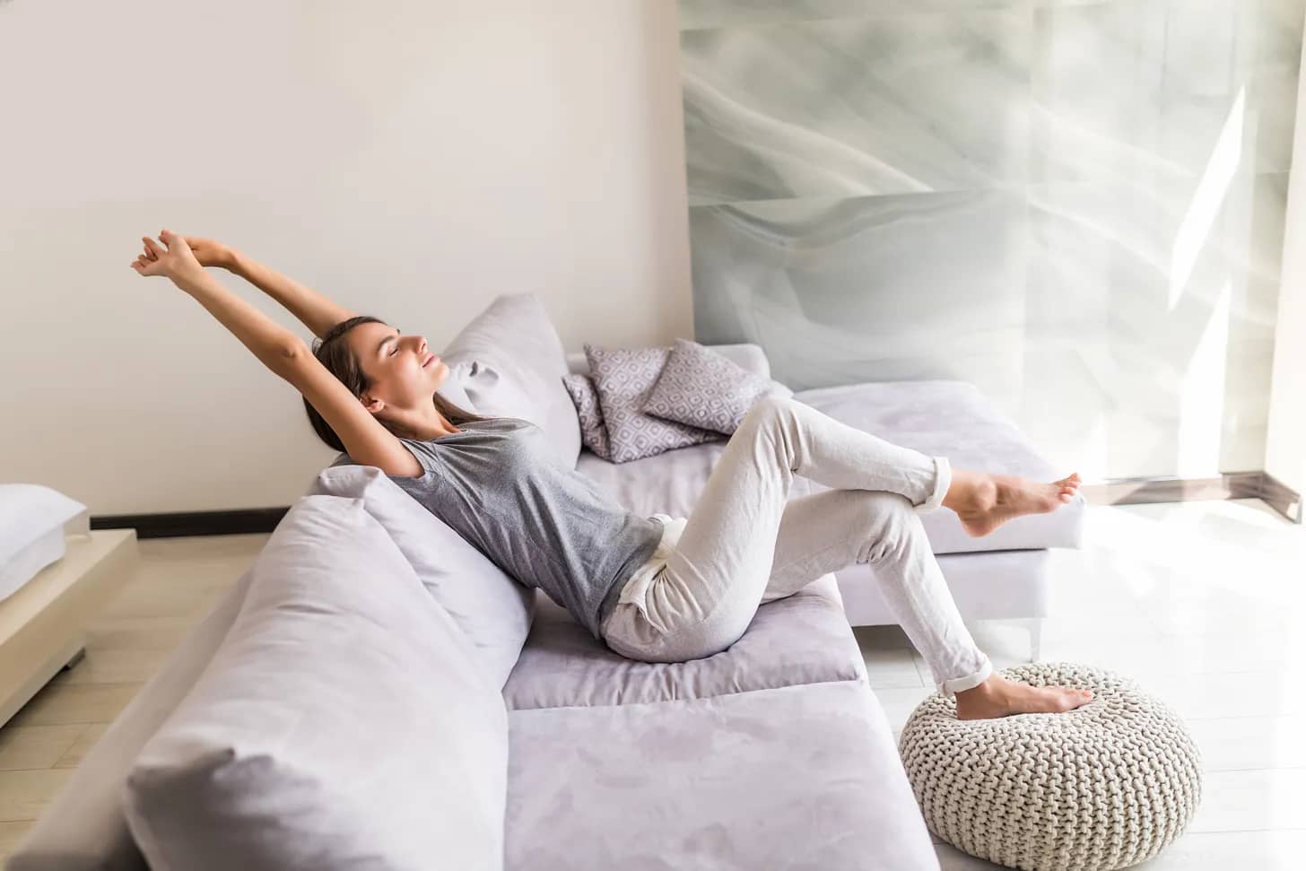 mulher jovem a relaxar e a alongar os braços, sentada num sofá dentro de casa.